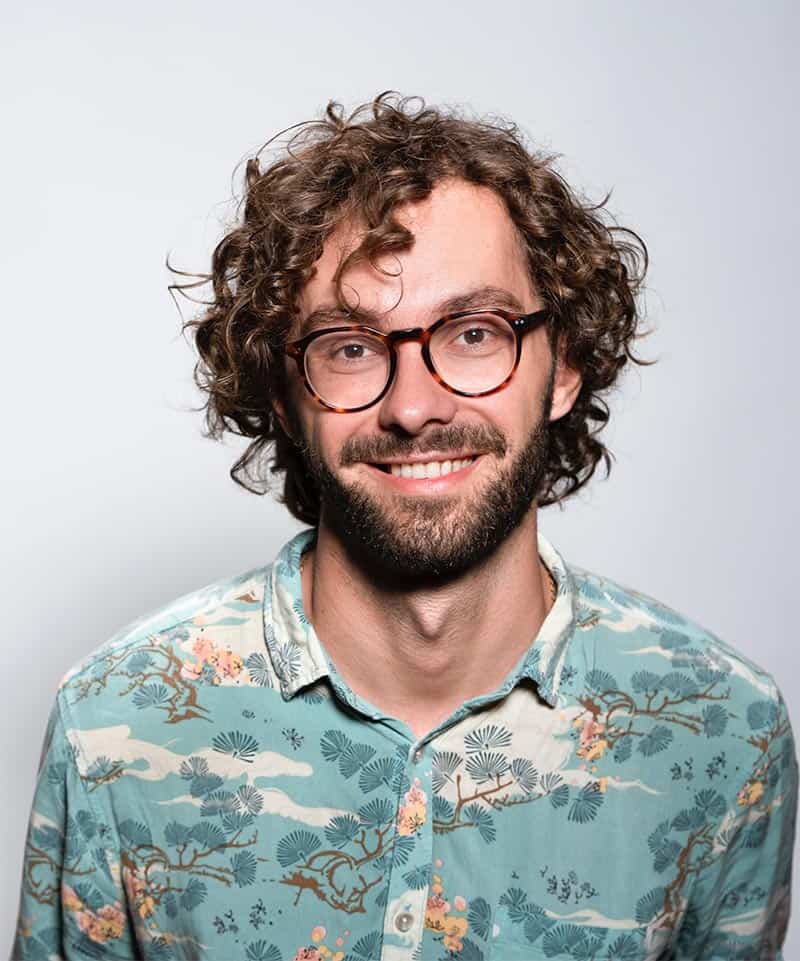 A man with curly hair, a beard, and glasses is smiling at the camera. He is wearing a light blue floral patterned shirt and standing against a plain light background.