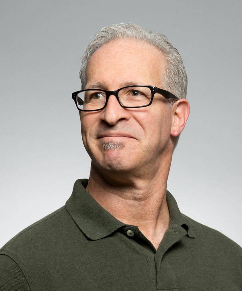 A middle-aged man with short gray hair, glasses, and a goatee, wearing a dark green polo shirt, looks slightly to the side against a plain gray background.