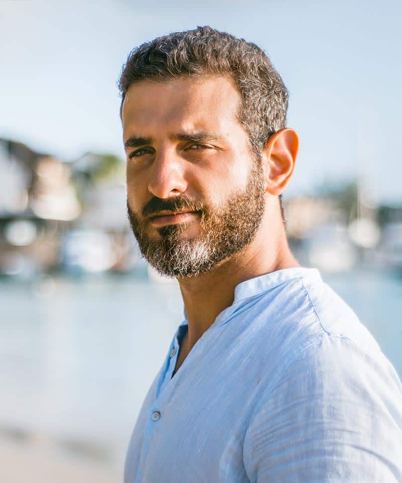A man with short curly hair and a beard, wearing a light blue shirt, stands outdoors with blurred water and buildings in the background.