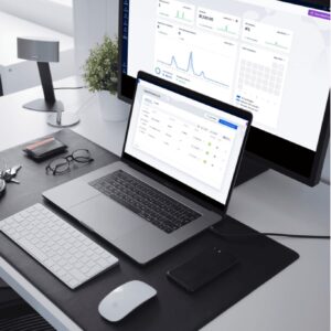 A modern desk setup with a laptop and external monitor showing data dashboards from the Professional Premium Plan, plus a keyboard, mouse, smartphone, glasses, and cardholder on a black desk mat.
