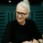 An older woman with gray hair in a bun and glasses sits at a table, smiling and holding a pen, with sticky notes and a device in front of her.