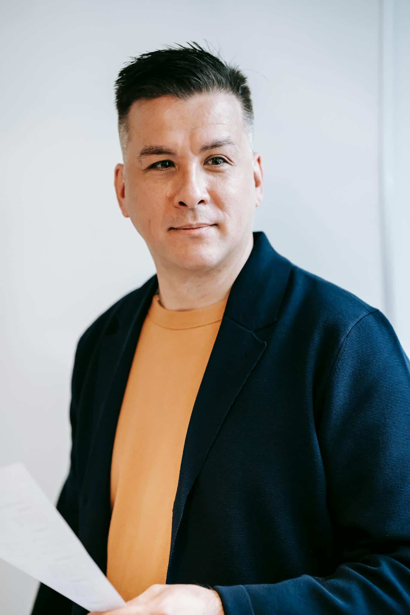 Man with short dark hair wearing a navy jacket and yellow shirt holds papers and looks to the side against a light background.