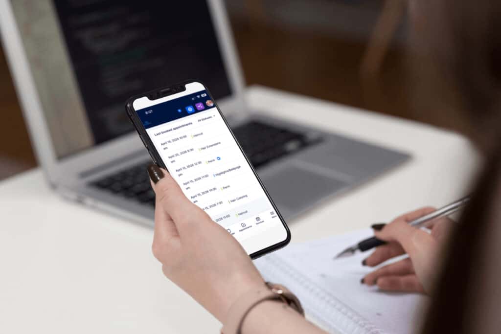 A person holds a smartphone displaying a mobile app with a transaction list while sitting at a desk with a laptop and writing in a notebook.