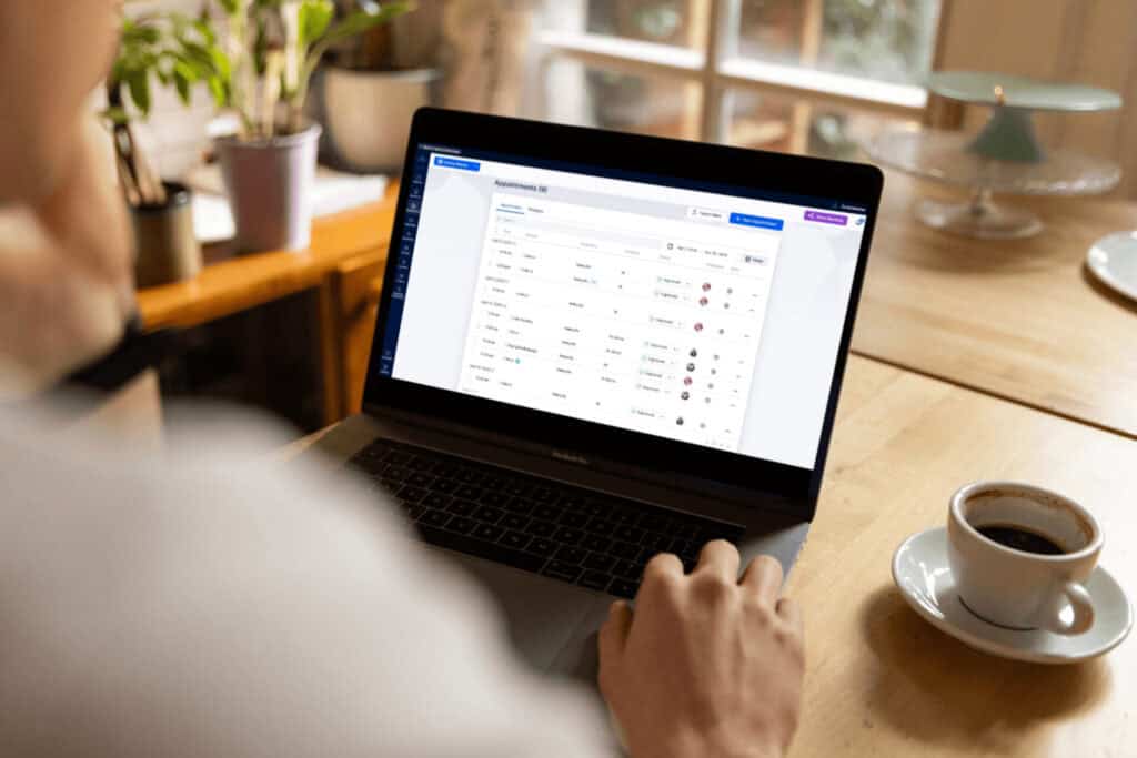 Person using a laptop at a table with a coffee cup; the screen displays a management dashboard with various data entries and status indicators.