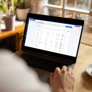 Person using a laptop at a table with a coffee cup; the screen displays a management dashboard with various data entries and status indicators.
