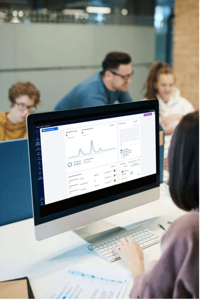 Person working at a computer displaying analytics software, with three colleagues having a discussion in the background in a modern office setting.