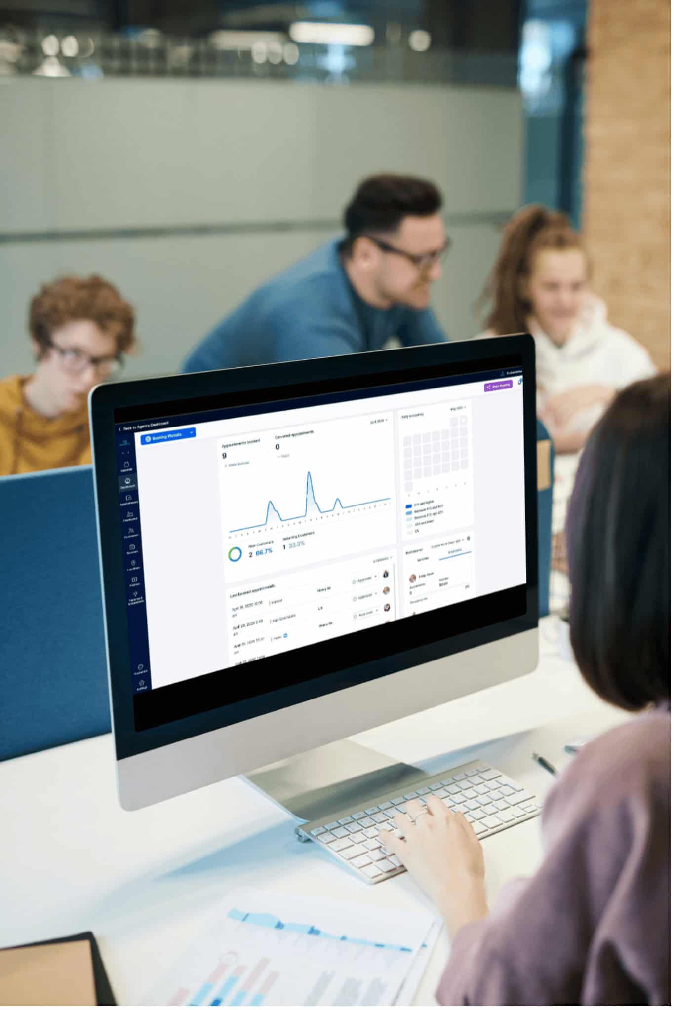 Person working at a computer displaying analytics software, with three colleagues having a discussion in the background in a modern office setting.