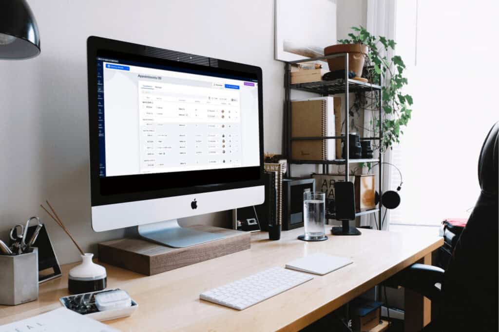 A desktop computer on a wooden desk displays an appointment management system; office supplies, a water glass, and a potted plant are also visible in a bright workspace.