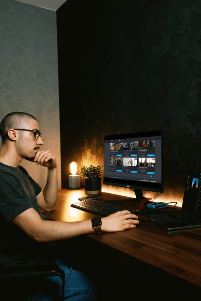 A man sits at a desk in a dimly lit room, looking at a computer monitor displaying various video thumbnails. A plant, lamp, and laptop are also on the desk.