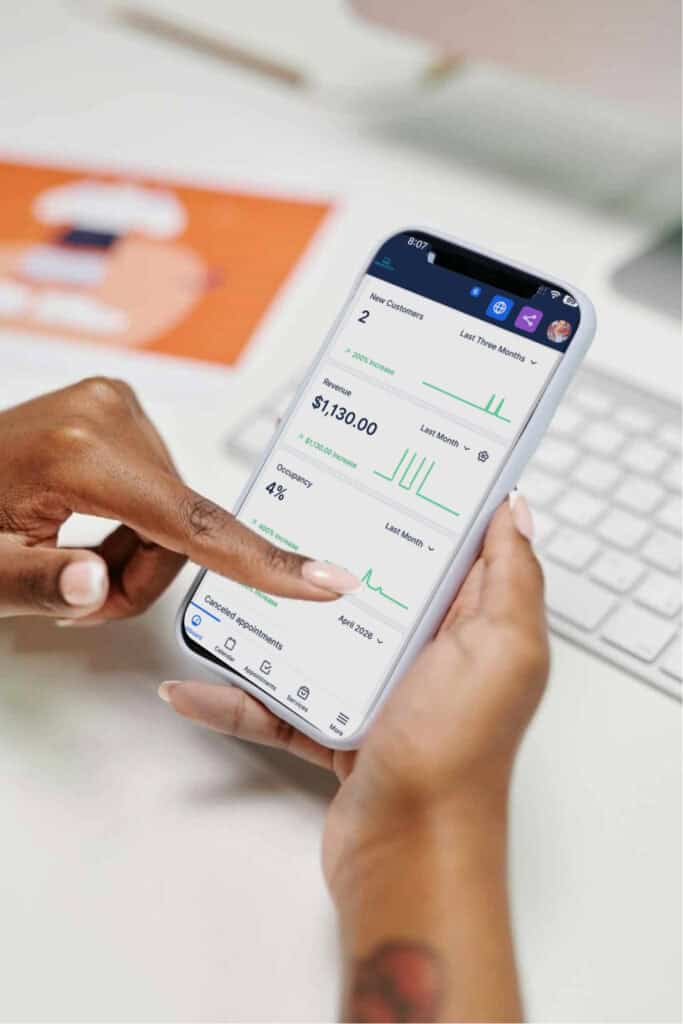 A person uses a smartphone displaying business analytics, with graphs and financial data visible on the screen, near a keyboard and document.
